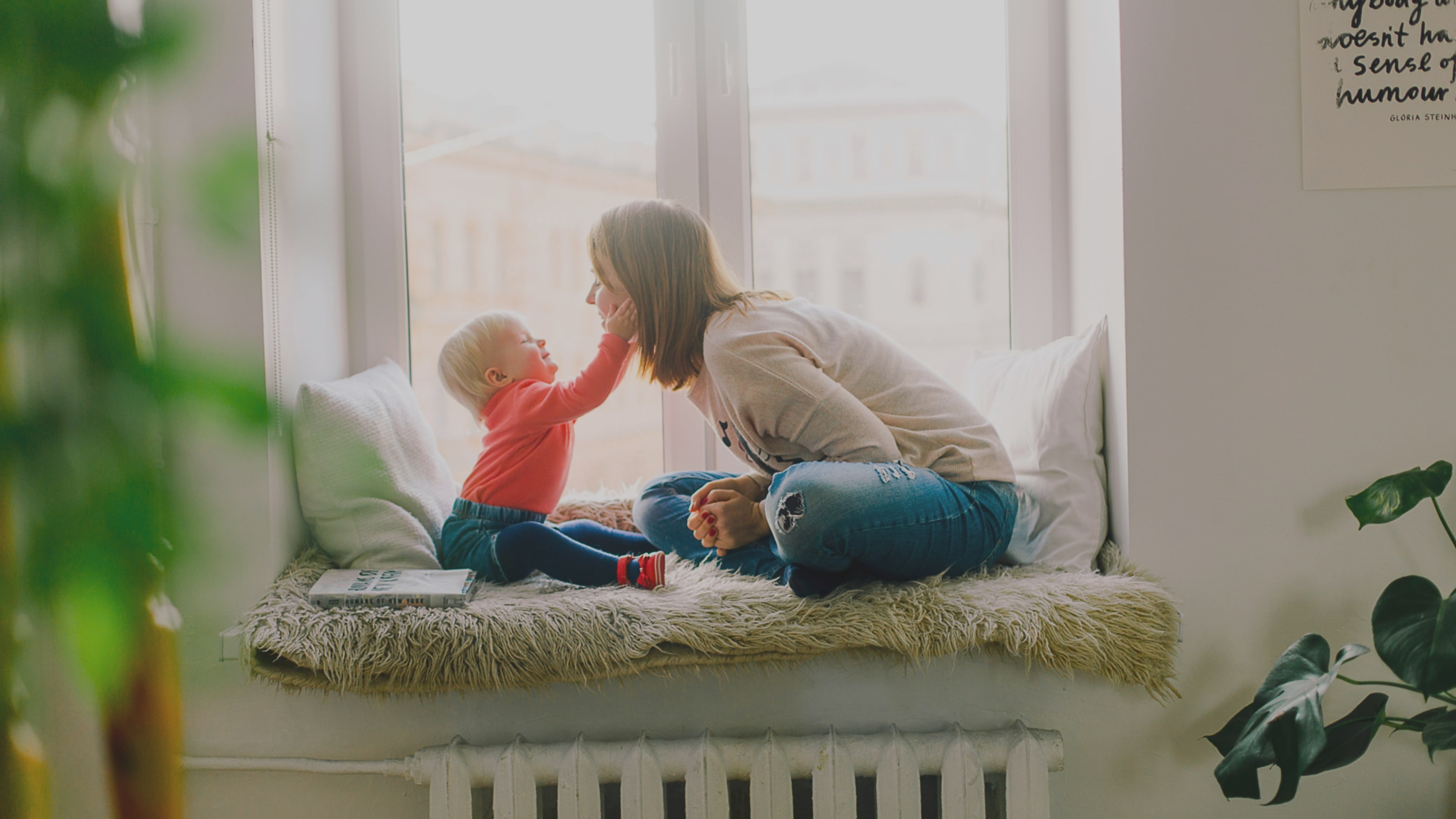 Baby affectionately touching mother’s face with its hands while they are sitting on a fluffy mat at a window seat