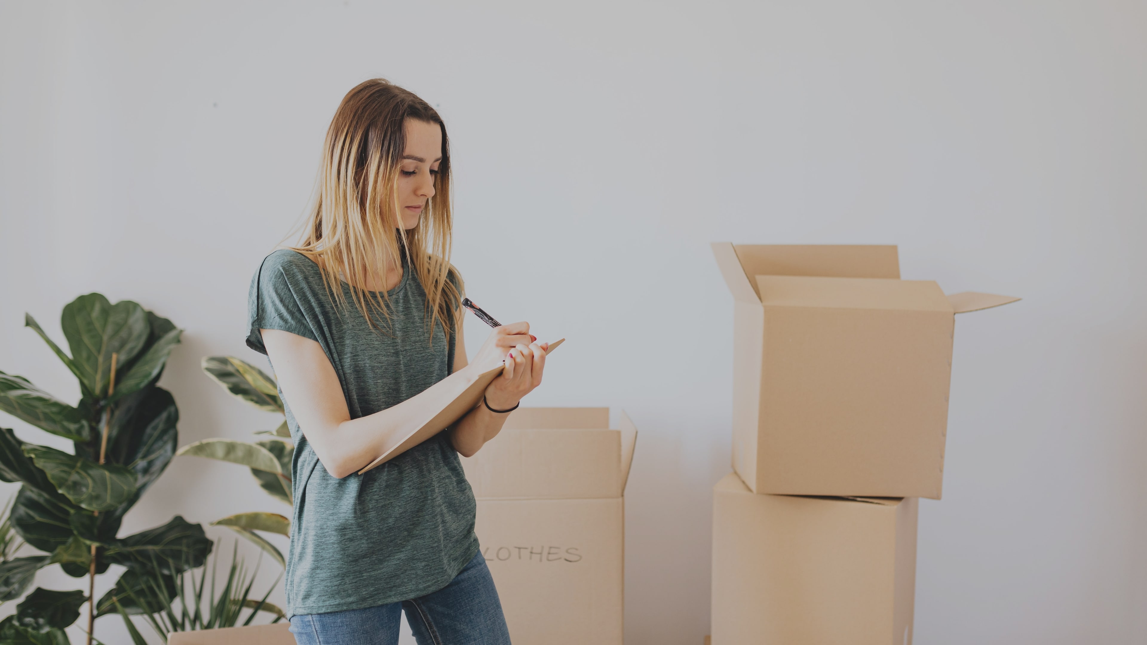 Homeowner standing beside a plant and cardboard boxes while making a list on her notebook